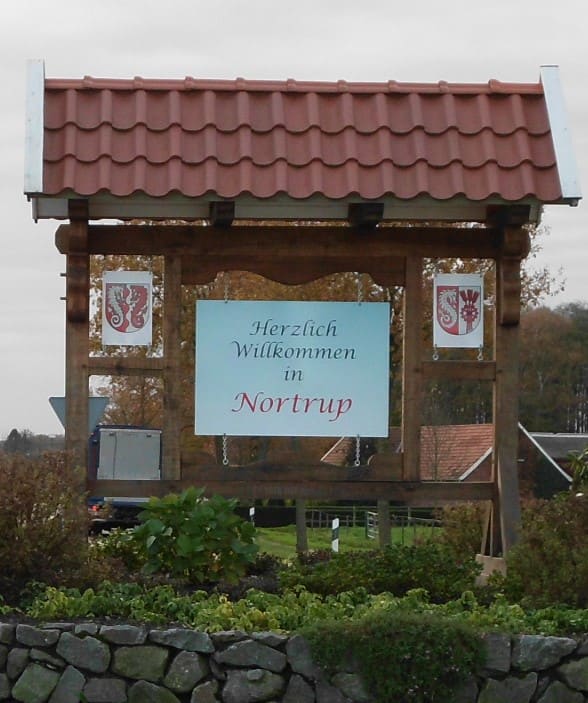 Überdachte Holztafel „Herzlich Willkommen in Nortrup“ mit zwei Wappen.
