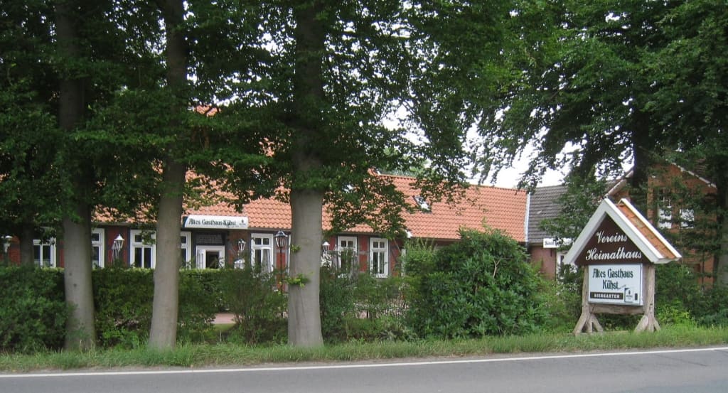Das Heimathaus hinter Bäumen an der Straße, davor ein Hinweisschild.